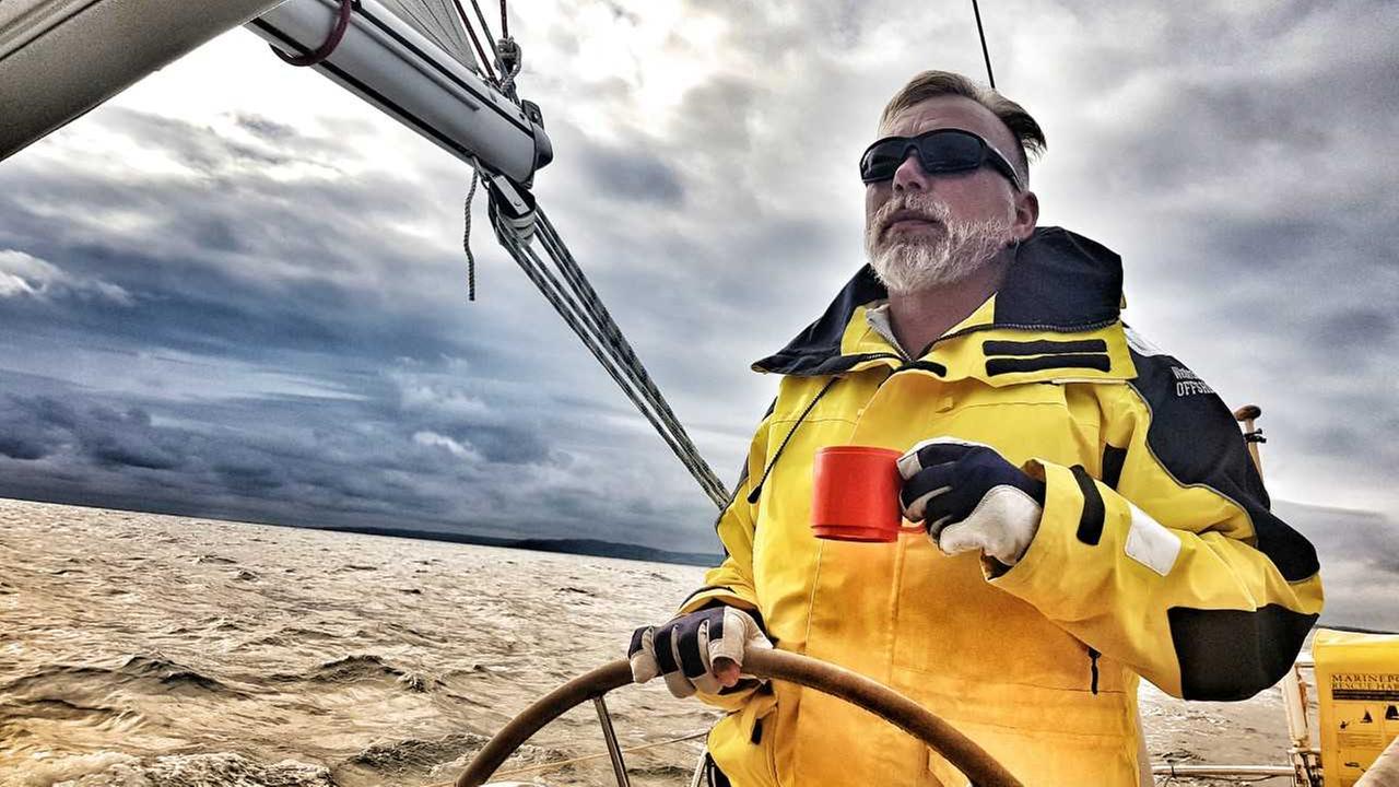 Thorsten Mohr trägt Handschuhe, eine Sonnenbrille und eine gelbe Jacke. Er steht am Ruder seines Segelbootes. In der Hand hält er eine rote Tasse. Am Himmel sind graue Wolken.