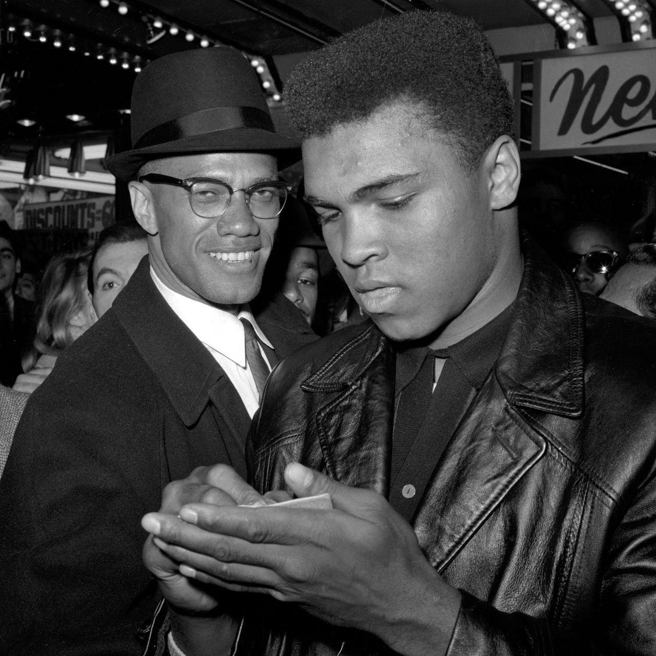 Der Boxer Muhammad Ali, in einer schwarzen Lederjacke, schreibt etwas auf einen Zettel in seiner linken Hand, hinter ihm steht der Bürgerrechtler Malcolm X in einem schwarzem Anzug und mit schwarzem Hut.