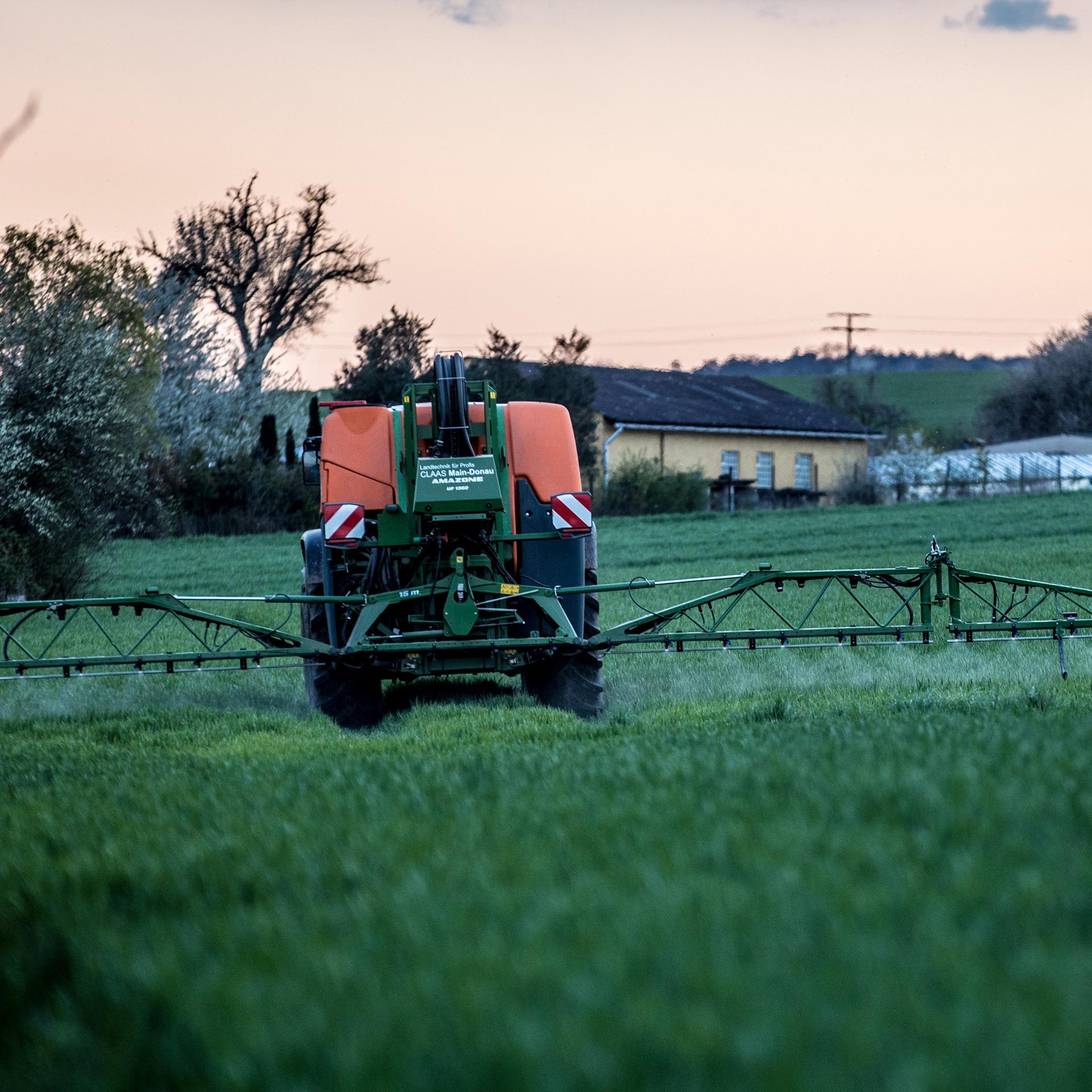 Ein Landwirt bringt mit seinem Traktor und einer großen Pflanzenschutzspritze Unkrautvernichter auf das Feld aus. Im Hintergrund ist ein Haus zu sehen. Es dämmert. 