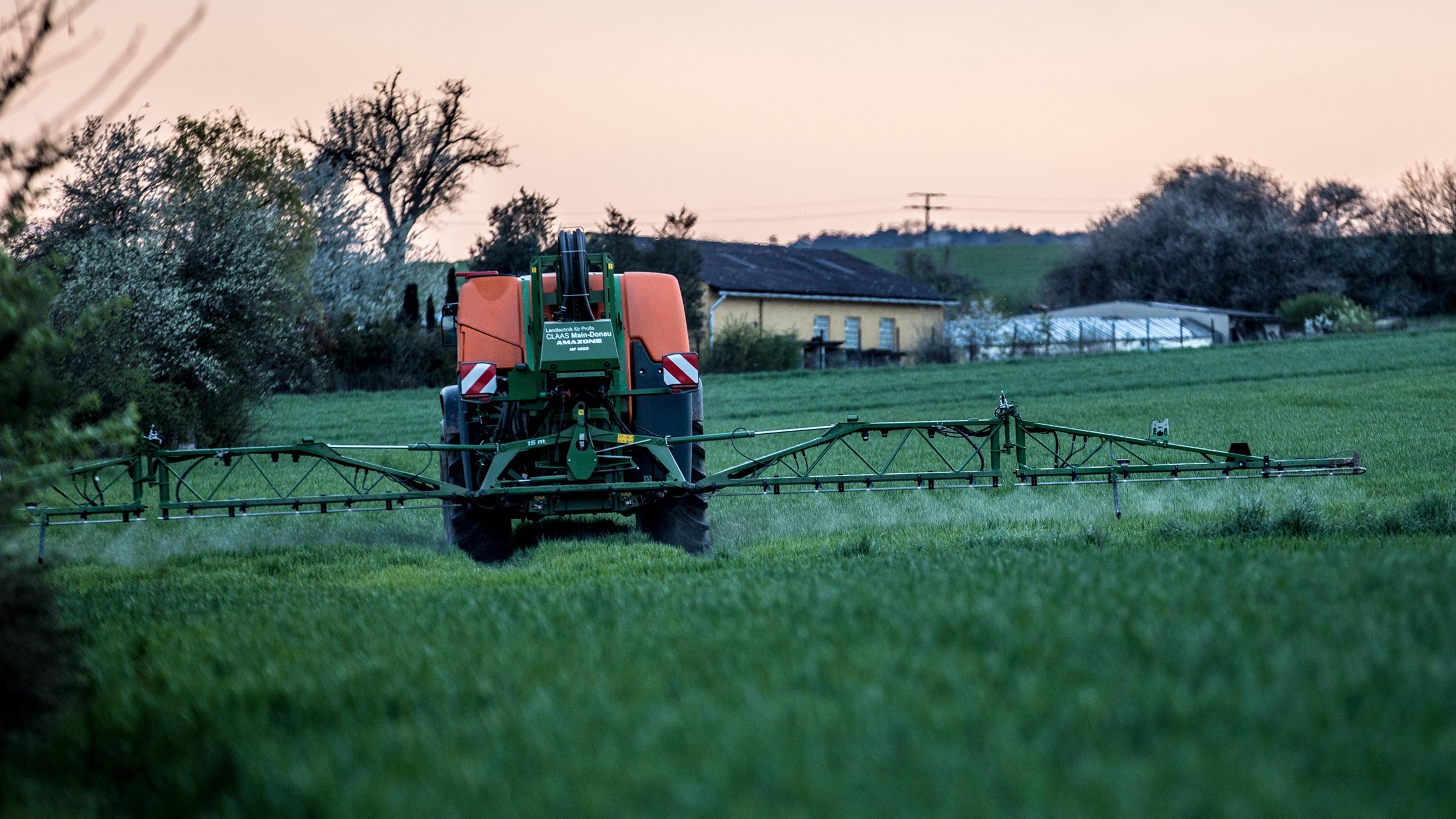 Ein Landwirt bringt mit seinem Traktor und einer großen Pflanzenschutzspritze Unkrautvernichter auf das Feld aus. Im Hintergrund ist ein Haus zu sehen. Es dämmert.