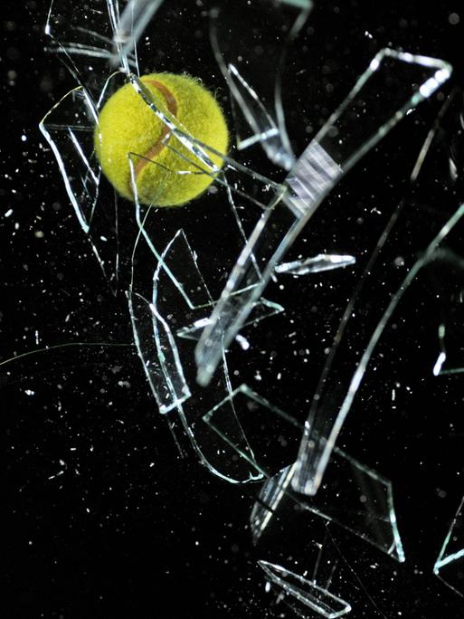 Ein Tennisball zerbricht eine Glasscheibe vor schwarzem Hintergrund, Glassplitter fliegen in alle Richtungen.