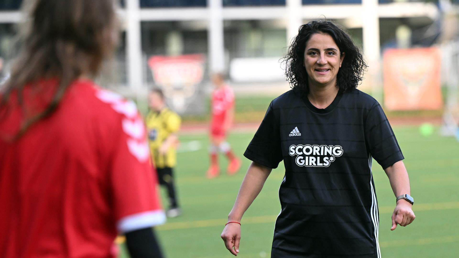 Tugba Tekkal steht mit "Scoring Girls" T-Shirt auf einem Sportplatz in Berlin und lächelt.
