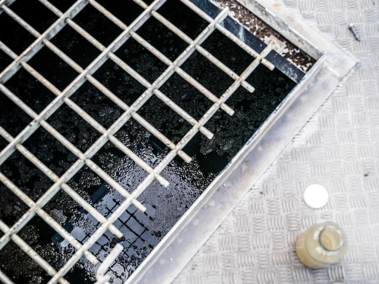 Abwasserprobe aus einer Kläranlage: Ein Glasbehälter mit Wasser steht neben einem großen Abfluss, in dessen Tiefe durch das Gitter dunkles Abwasser zu sehen ist. 