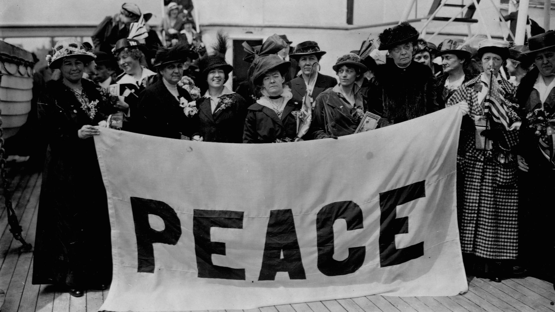 Historisches Schwarzweißfoto von Frauen, die 1915 auf einem Schiffsdeck stehen und ein Plakat mit der Aufschrift "Peace" in die Kameras halten.