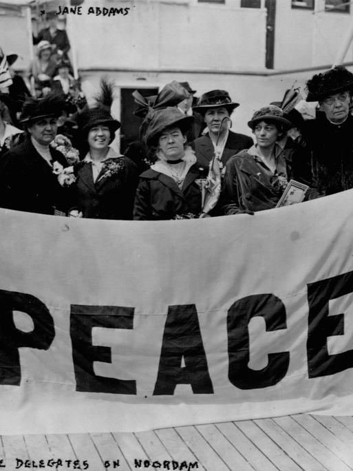 Historisches Schwarzweißfoto von Frauen, die 1915 auf einem Schiffsdeck stehen und ein Plakat mit der Aufschrift "Peace" in die Kameras halten.