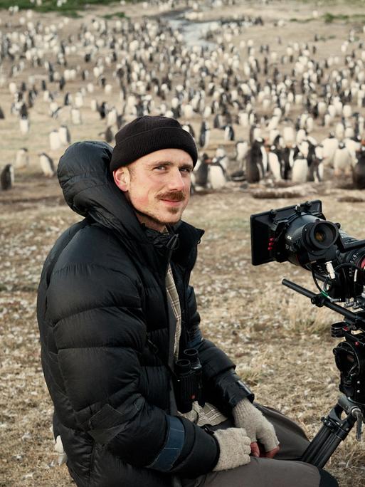 Rolf Steinmann steht mit seiner Kamera in der Steppe und lächelt in die Kamera. Im Hintergrund sind Pinguine zu sehen.