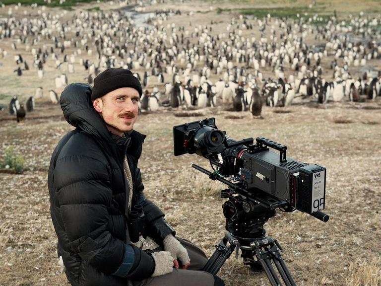 Rolf Steinmann steht mit seiner Kamera in der Steppe und lächelt in die Kamera. Im Hintergrund sind Pinguine zu sehen.