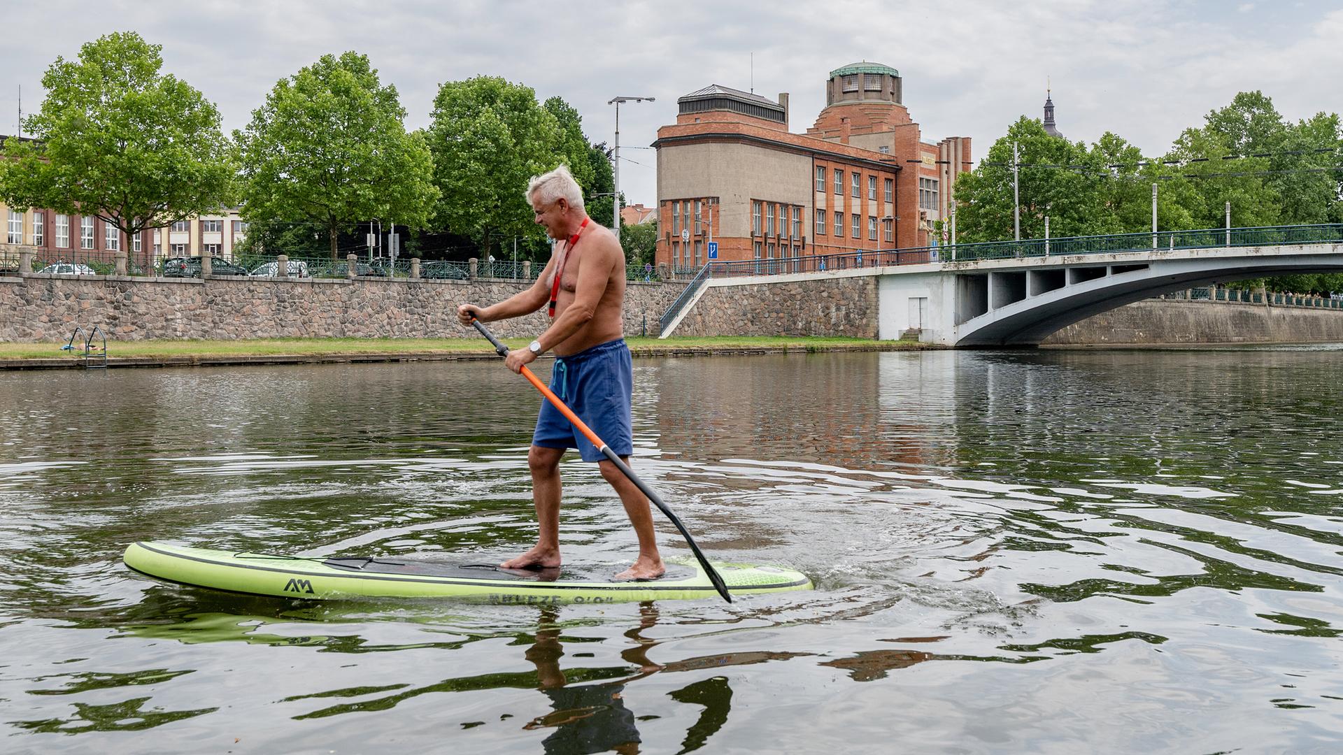 Ein weißhaariger Mann paddelt im Stehen auf einem SUP-Board auf der Elbe im Zentrum von Hradec Králové in der Tschechischen Republik Ein weißhaariger Mann paddelt im Stehen auf einem SUP-Board auf der Elbe im Zentrum von Hradec Králové in der Tschechischen Republik
