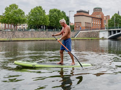 Ein weißhaariger Mann paddelt im Stehen auf einem SUP-Board auf der Elbe im Zentrum von Hradec Králové in der Tschechischen Republik