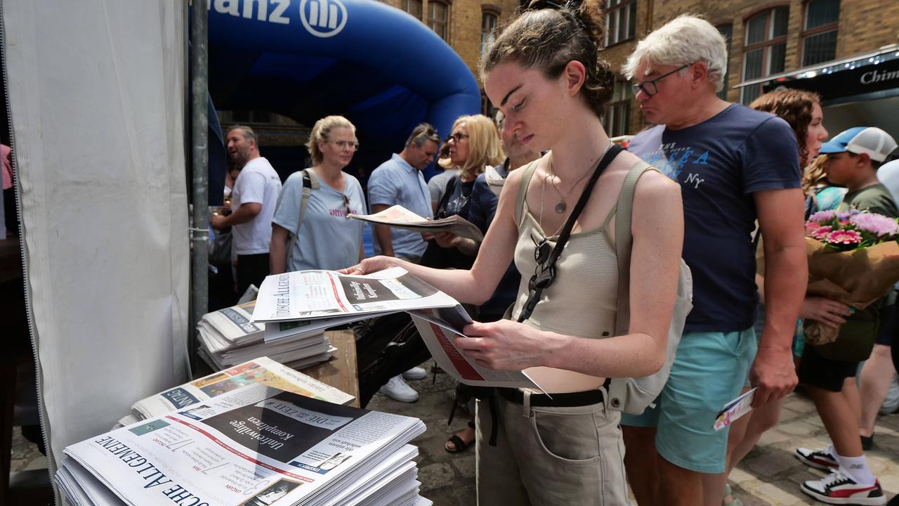 Eine sommerlich gekleidete Frau steht in einem Innenhof und nimmt ein Exemplar von einem Zeitungsstapel. Dahinter weitere Personen. 