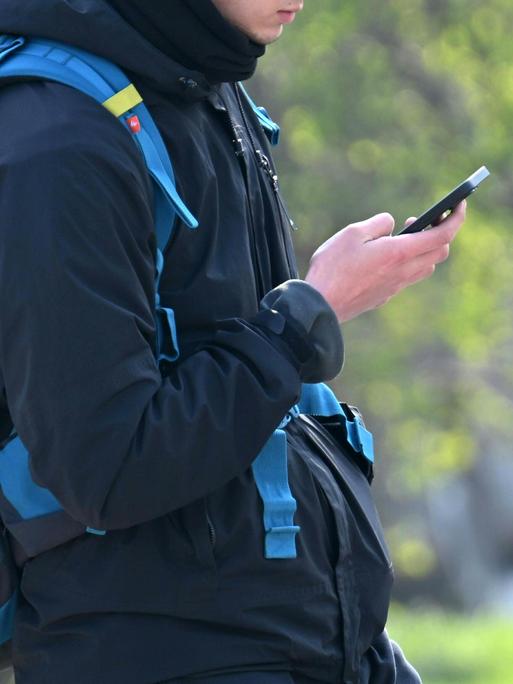 Ein Jugendlicher blickt auf ein Smartphone und trägt einen Schulranzen.