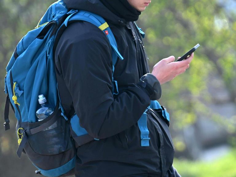 Ein Jugendlicher blickt auf ein Smartphone und trägt einen Schulranzen.