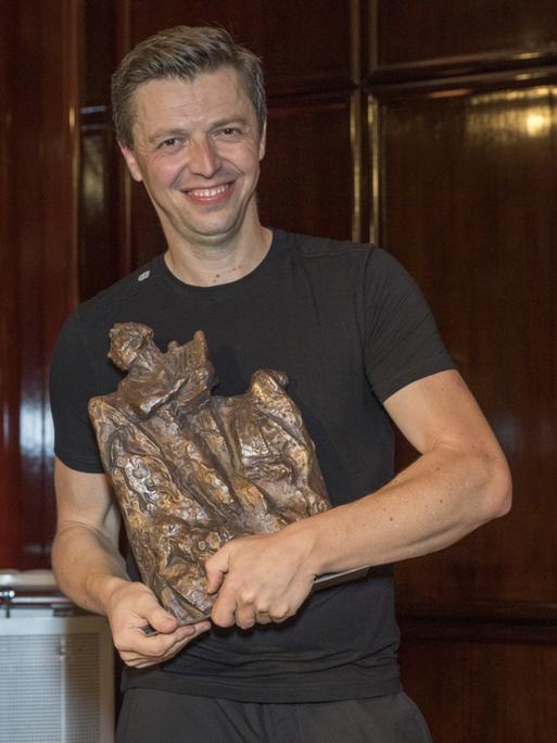 Ein junger Mann mit kurzem, dunkelblondem Haar steht in schwarzem T-Shirt vor einer Holzwand. In den Händen hält er eine große Bronze-Skulptur.