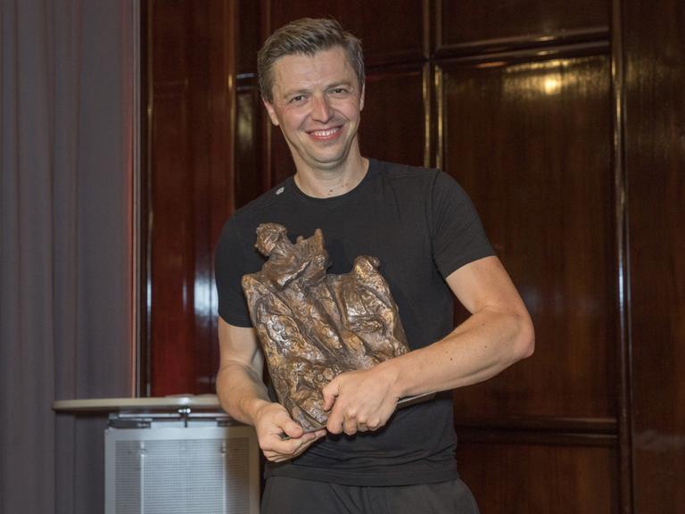 Ein junger Mann mit kurzem, dunkelblondem Haar steht in schwarzem T-Shirt vor einer Holzwand. In den Händen hält er eine große Bronze-Skulptur.