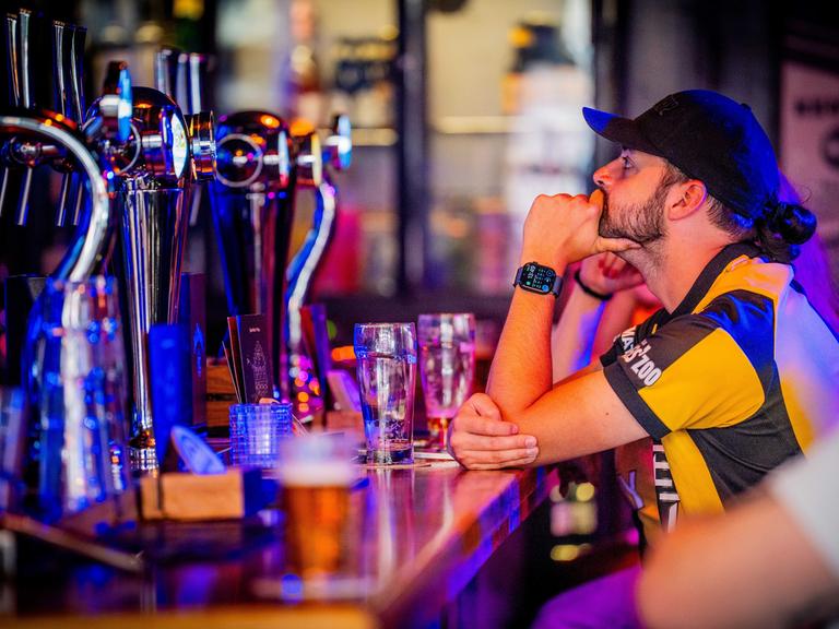 Ein Fußballfan im gelben Trikot sitzt an einer Theke. Er schaut nach links oben, wahrscheinlich auf einen Bildschirm. Links im Bild ist unscharf die Zapfanlage zu sehen.