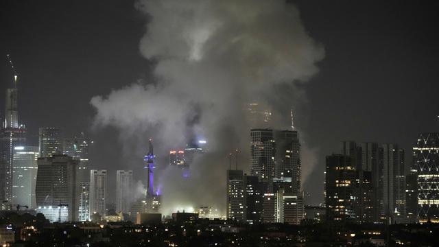Es ist Nacht. Man sieht Gebäude und die Lichter Tel Avivs aus der Ferne. Dichter grauer Rauch steigt über Häusern auf.