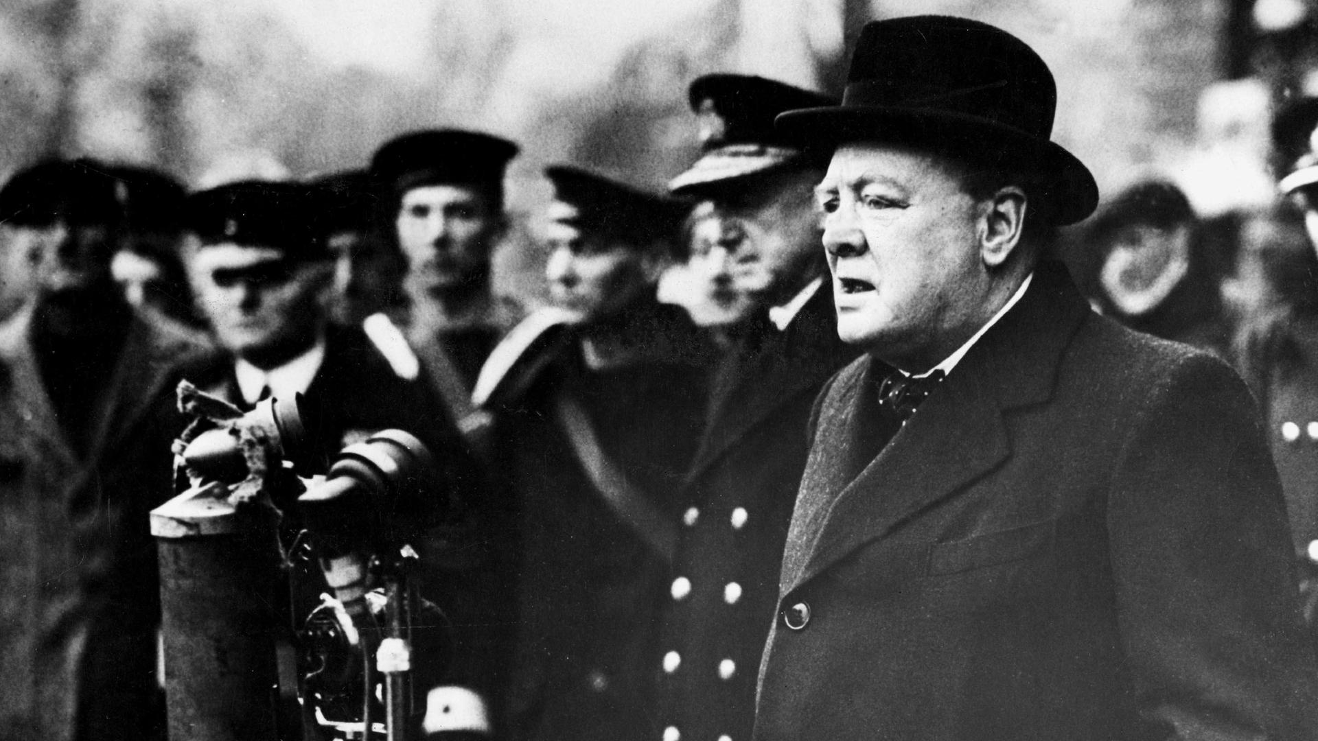 Winston Churchill steht an mehreren Mikrofonen und spricht zu Marine-Soldaten, die im Hintergrund zu sehen sind. 