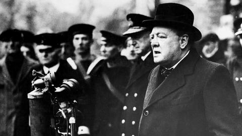 Winston Churchill steht an mehreren Mikrofonen und spricht zu Marine-Soldaten, die im Hintergrund zu sehen sind. 
