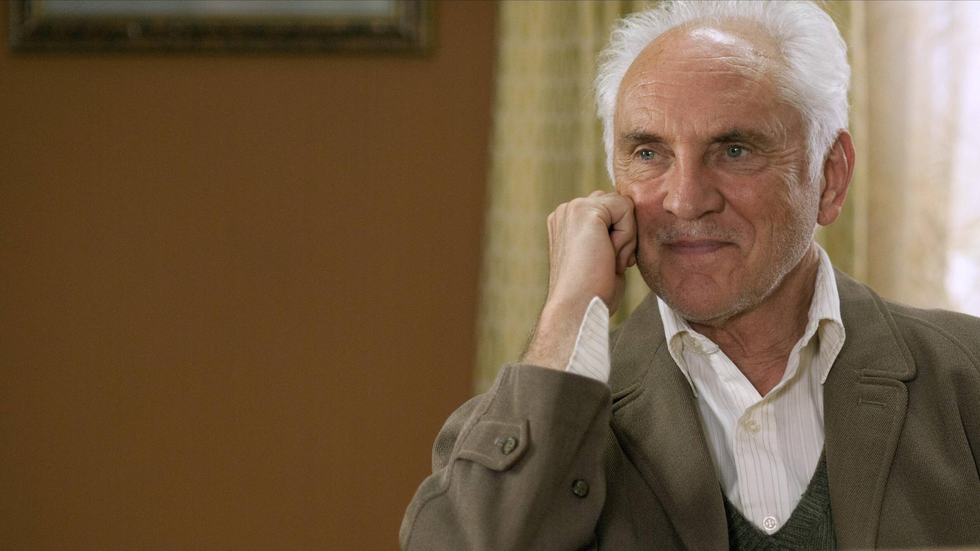 Das Bild zeigt einen freundlich lächelnden Terence Stamp, der seinen Kopf an seine rechte Hand anlehnt. Seine Haare sind weiß.