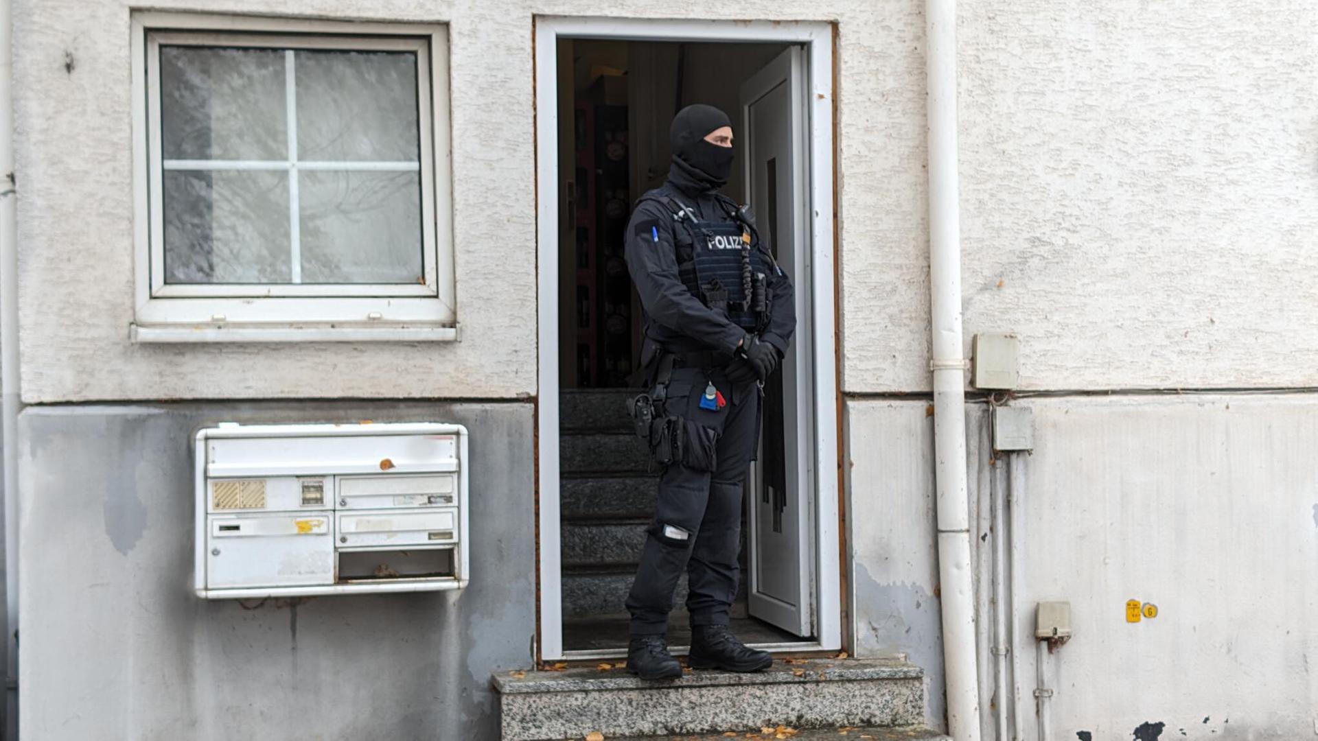 Ein Polizist in schwarzer Kampfmontur und mit hochgezogener Sturmhaube steht vor der Eingangstür eines weißgetünchten Wohnhauses. An der Wand hängt eine Briefkasten-Anlage.