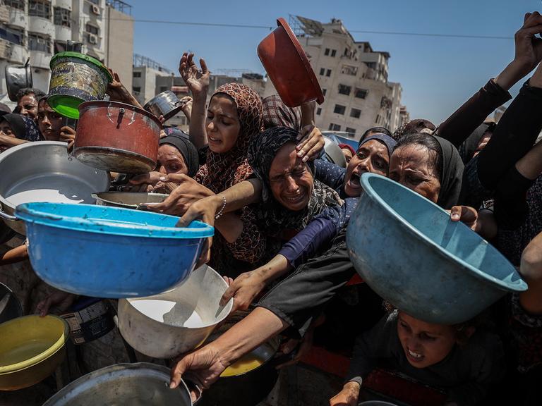 Frauen, Männer und Kinder in Gaza-Stadt strecken Helfer, die Lebensmittel verteilen, bunte Plastikschüsseln entgegen.