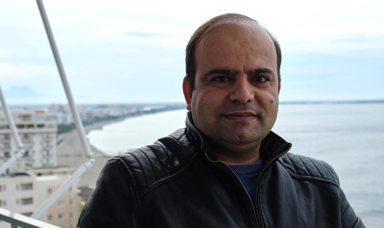 Faisal Karimi trägt eine schwarze Lederjacke. Hinter ihm ist das Meer und der Strand zu sehen.