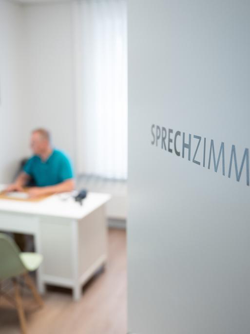 Ein Hausarzt sitzt in einem Sprechzimmer seiner Hausarztpraxis an einem Schreibtisch und arbeitet am Computer. Ein Hausarzt sitzt in einem Sprechzimmer seiner Hausarztpraxis an einem Schreibtisch und arbeitet am Computer.