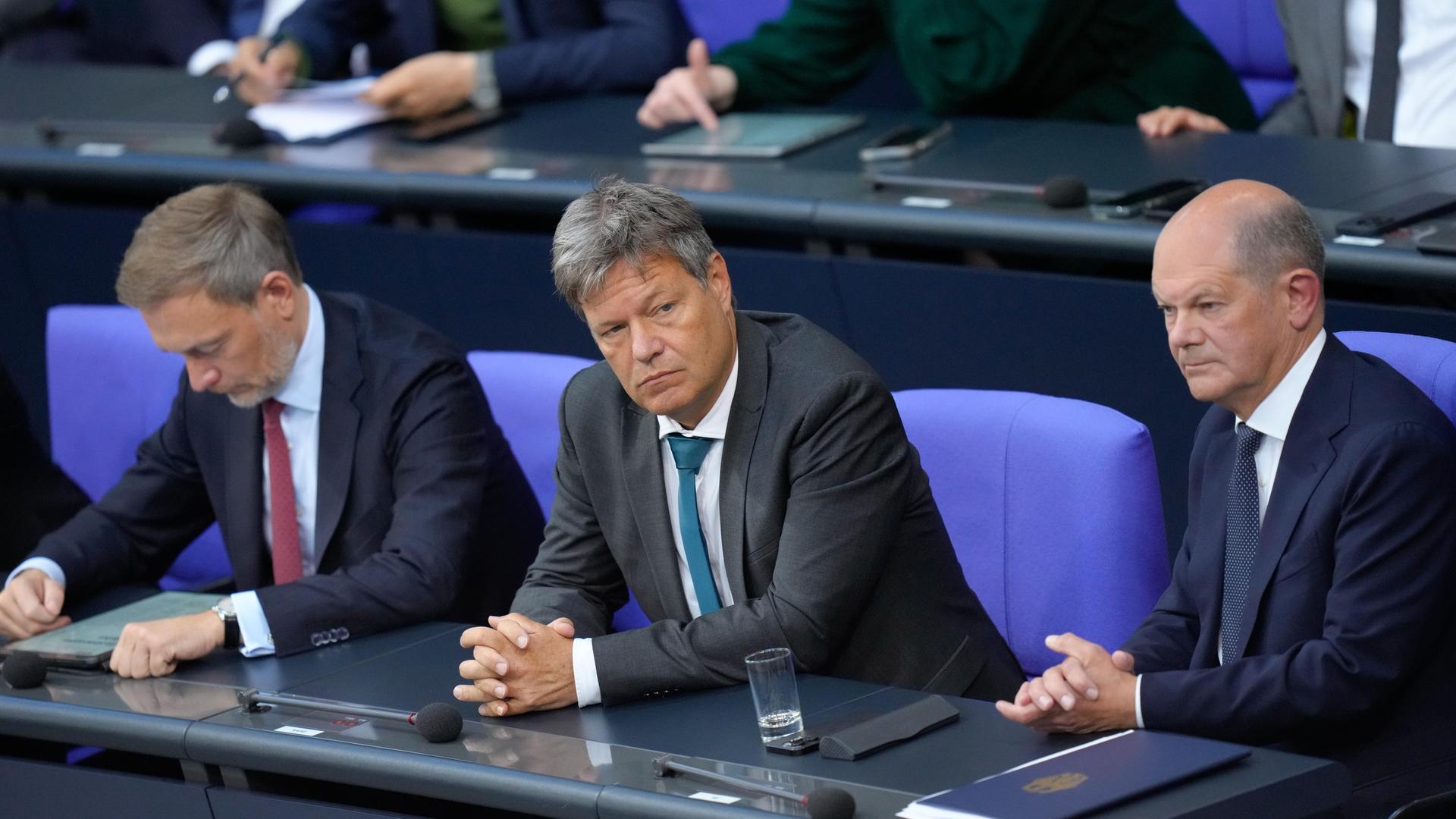 Bundeskanzler Olaf Scholz, Wirtschaftsminister Robert Habeck und Finanzminister Christian Lindner sitzen nebeneinander auf der Regierungsbank im Bundestag und schauen gelangweilt.