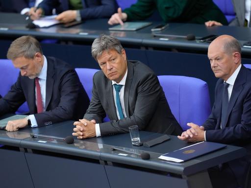 Bundeskanzler Olaf Scholz, Wirtschaftsminister Robert Habeck und Finanzminister Christian Lindner sitzen nebeneinander auf der Regierungsbank im Bundestag und schauen gelangweilt.