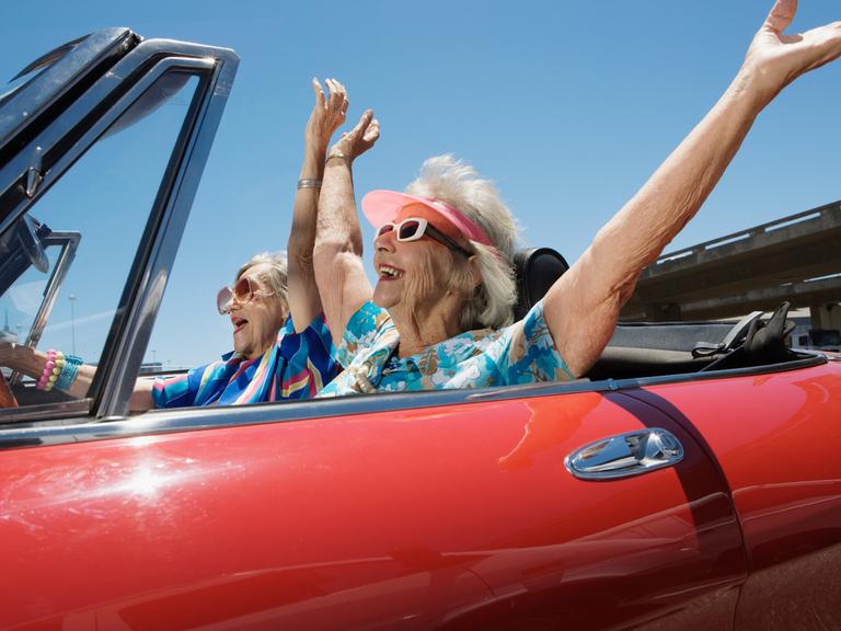 Zwei glückliche ältere Frauen mit ausgestreckten Armen in einem roten Cabrio.