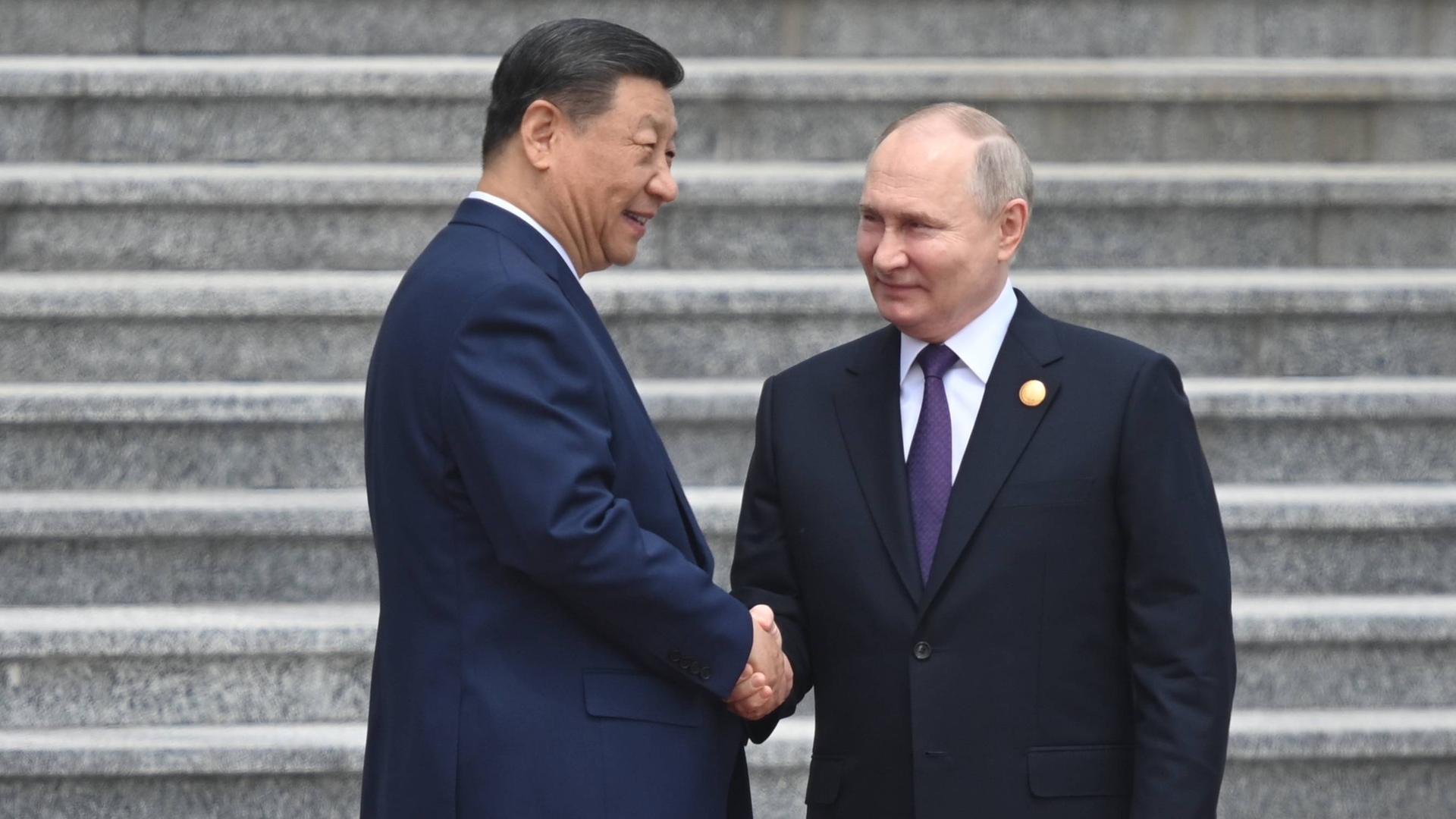 Der russische Präsident Vladimir Putin und der chinesische Präsident Xi Jinping schütteln sich die Hand. Sie stehen auf einer grauen Treppe.