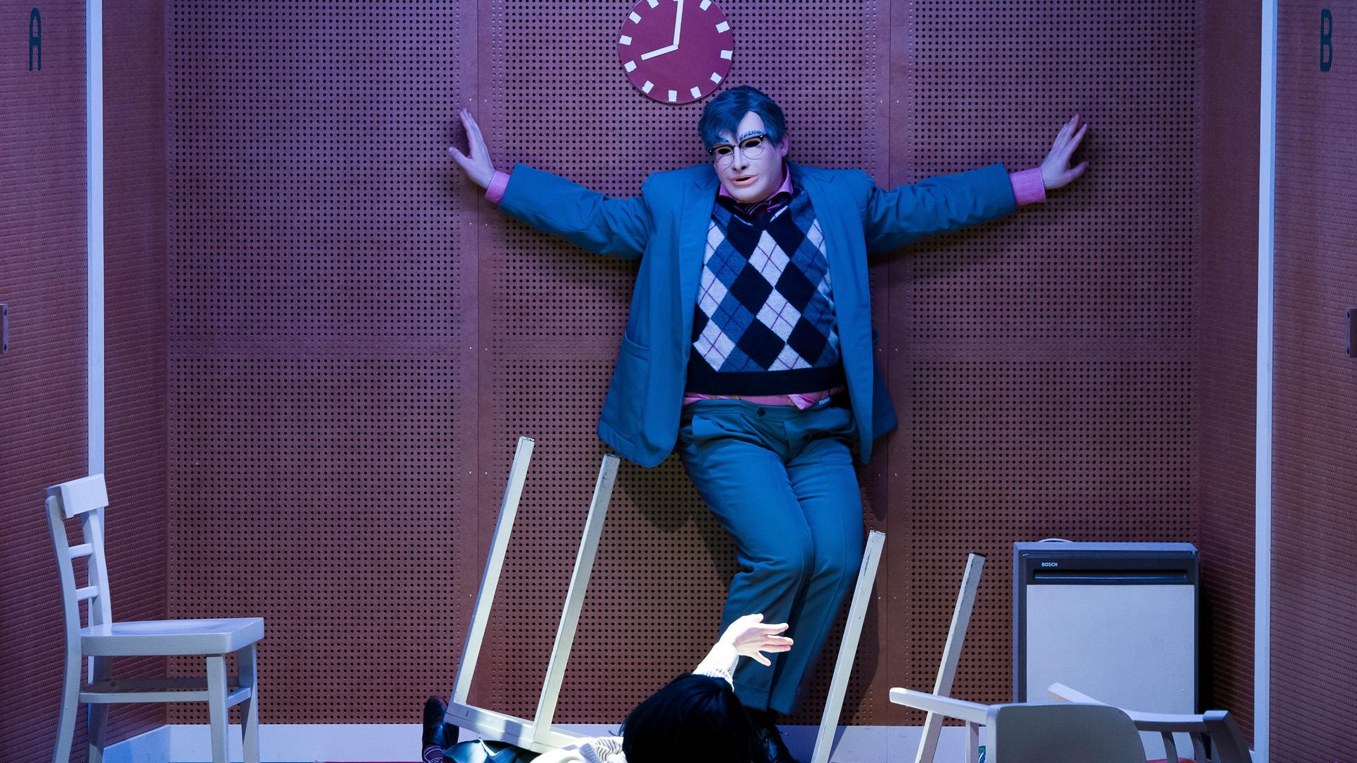 Szene aus dem Theaterstück "humanistää!" am Volkstheater in Wien. Eine Person mit ausgebreiteten Armen und mit Maske steht in einem grauen Anzug mit kariertem Pullunder vor einer braunen Wand.