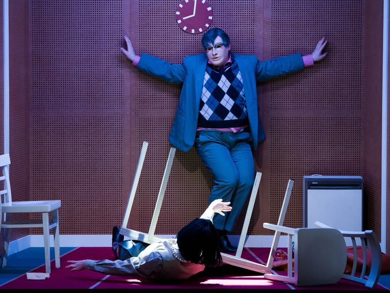 Szene aus dem Theaterstück "humanistää!" am Volkstheater in Wien. Eine Person mit ausgebreiteten Armen und mit Maske steht in einem grauen Anzug mit kariertem Pullunder vor einer braunen Wand.