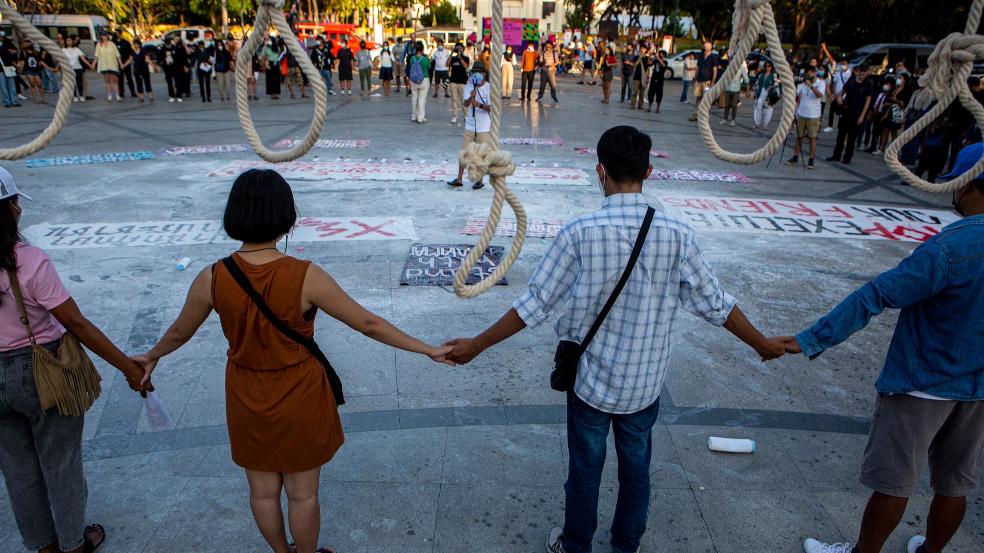 Demonstranten stehen vor sieben symbolisch aufgehängten Galgen und fassen einander an den Händen.