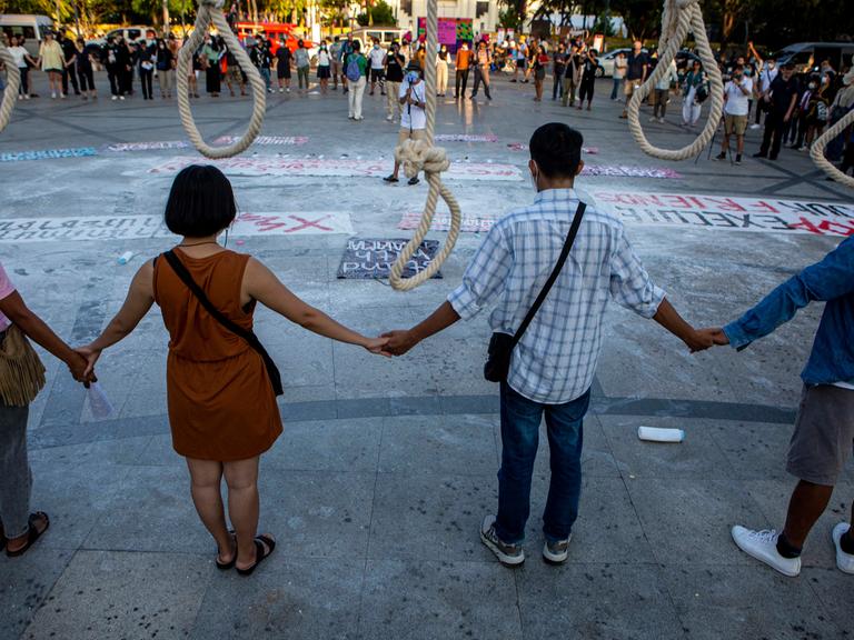 Demonstranten stehen vor sieben symbolisch aufgehängten Galgen und fassen einander an den Händen.