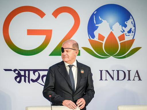 Bundeskanzler Olaf Scholz (SPD) steht mit Augenklappe vor dem Logo des G20-Gipfels in Neu Delhi in Indien.