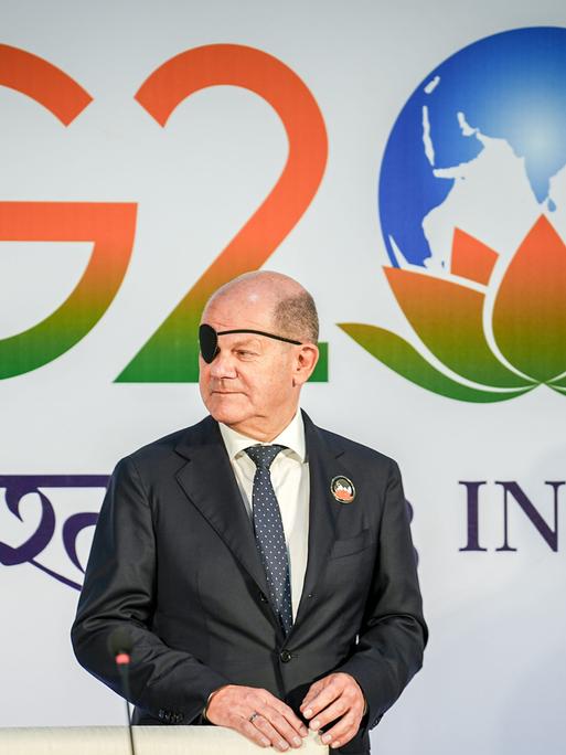 Bundeskanzler Olaf Scholz (SPD) steht mit Augenklappe vor dem Logo des G20-Gipfels in Neu Delhi in Indien.