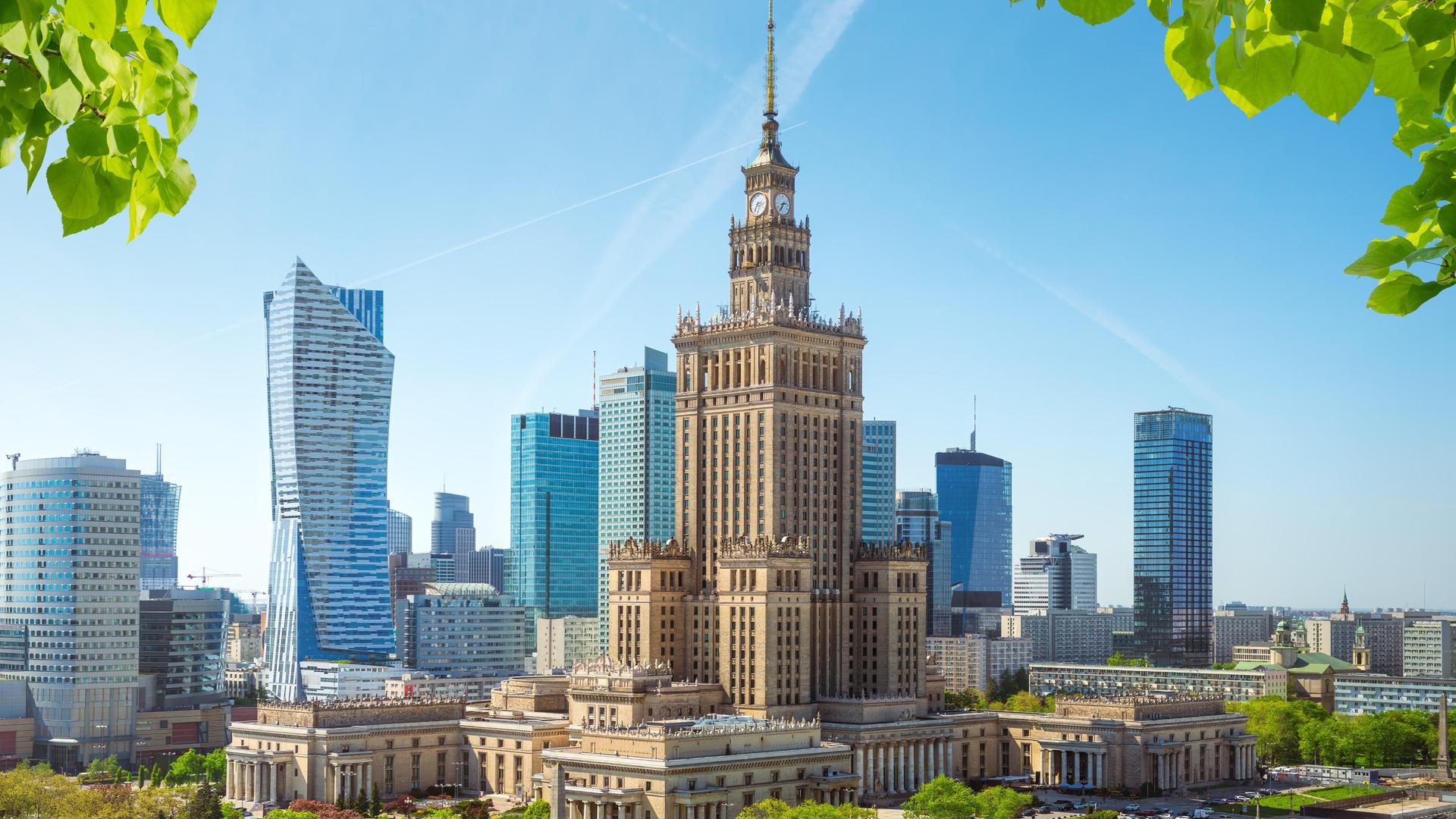 Der Kulturpalast in Polen, von einer Anhöhe aus betrachtet. Am Bildrand sind Blätter zu sehen, hinter dem zweithöchsten Gebäude Polens ist die moderne Silhouette Warschaus mit Wolkenkratzern zu sehen.