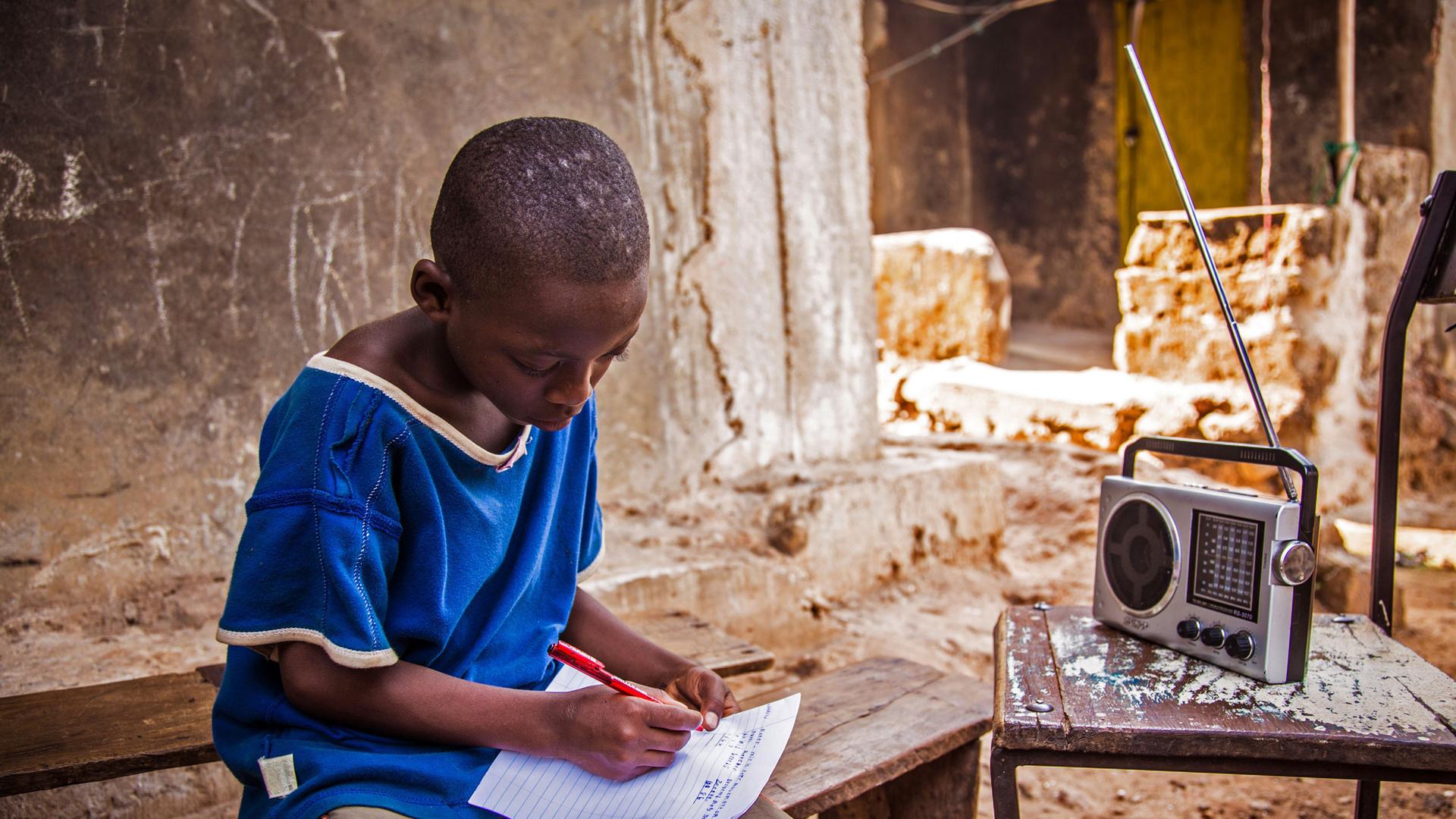 Ein junger sitzt in Sierra Leone mit Stift und Papier vor dem Radiogerät und erledigt seine Schulaufgaben.