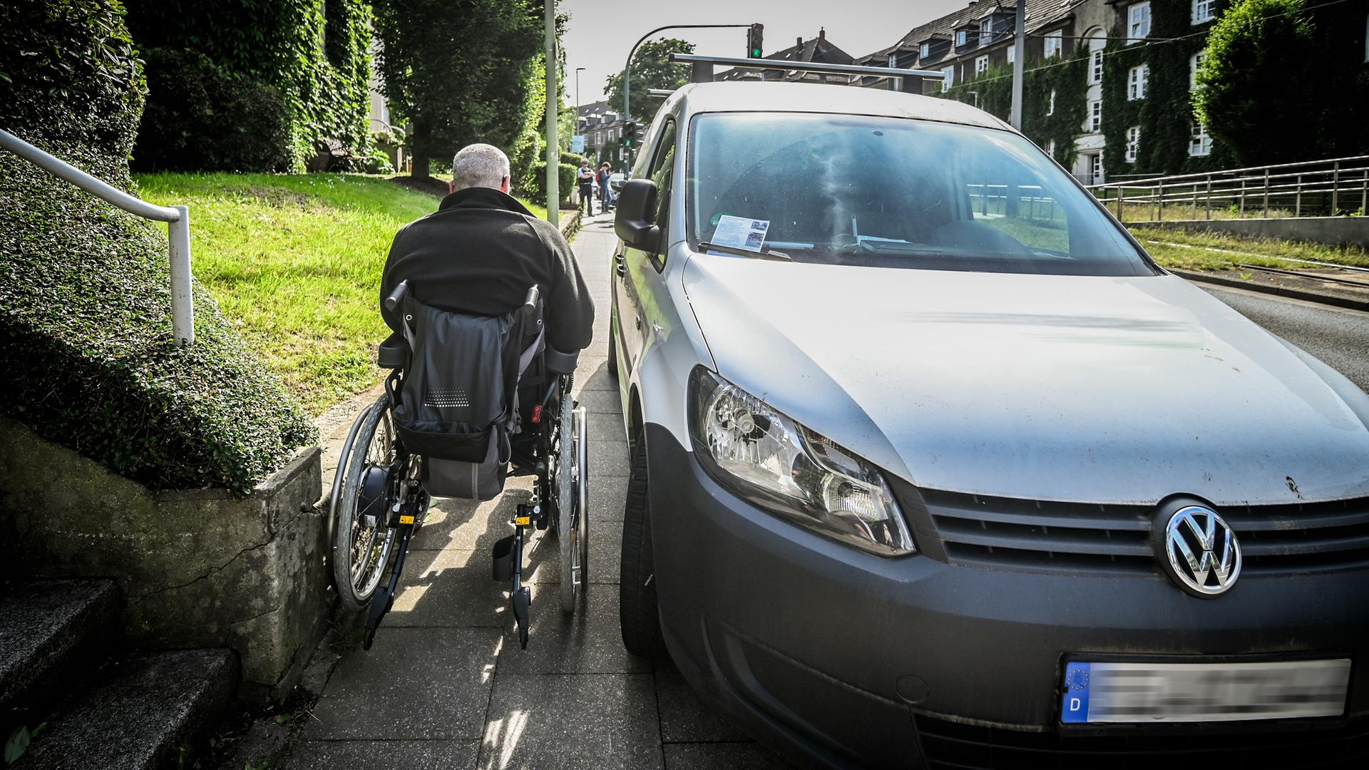 Ein Mann im einem Roll-Stuhl benutzt den Geh-Weg. Ein Auto parkt auf dem Geh-Weg. Der Mann hat daher nicht viel Platz.