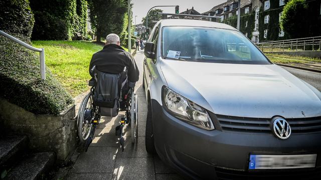Ein Mann im einem Roll-Stuhl benutzt den Geh-Weg. Ein Auto parkt auf dem Geh-Weg. Der Mann hat daher nicht viel Platz.