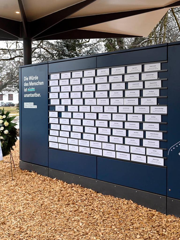 Auf einer dunkelblauen Wand hängen weiße Schilder mit den Namen der Opfer der nationalsozialistischen Euthanasie-Verbrechen. Daneben steht ein Blumenkranz.
