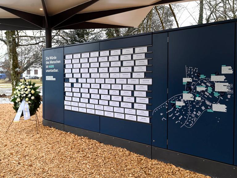 Auf einer dunkelblauen Wand hängen weiße Schilder mit den Namen der Opfer der nationalsozialistischen Euthanasie-Verbrechen. Daneben steht ein Blumenkranz.
