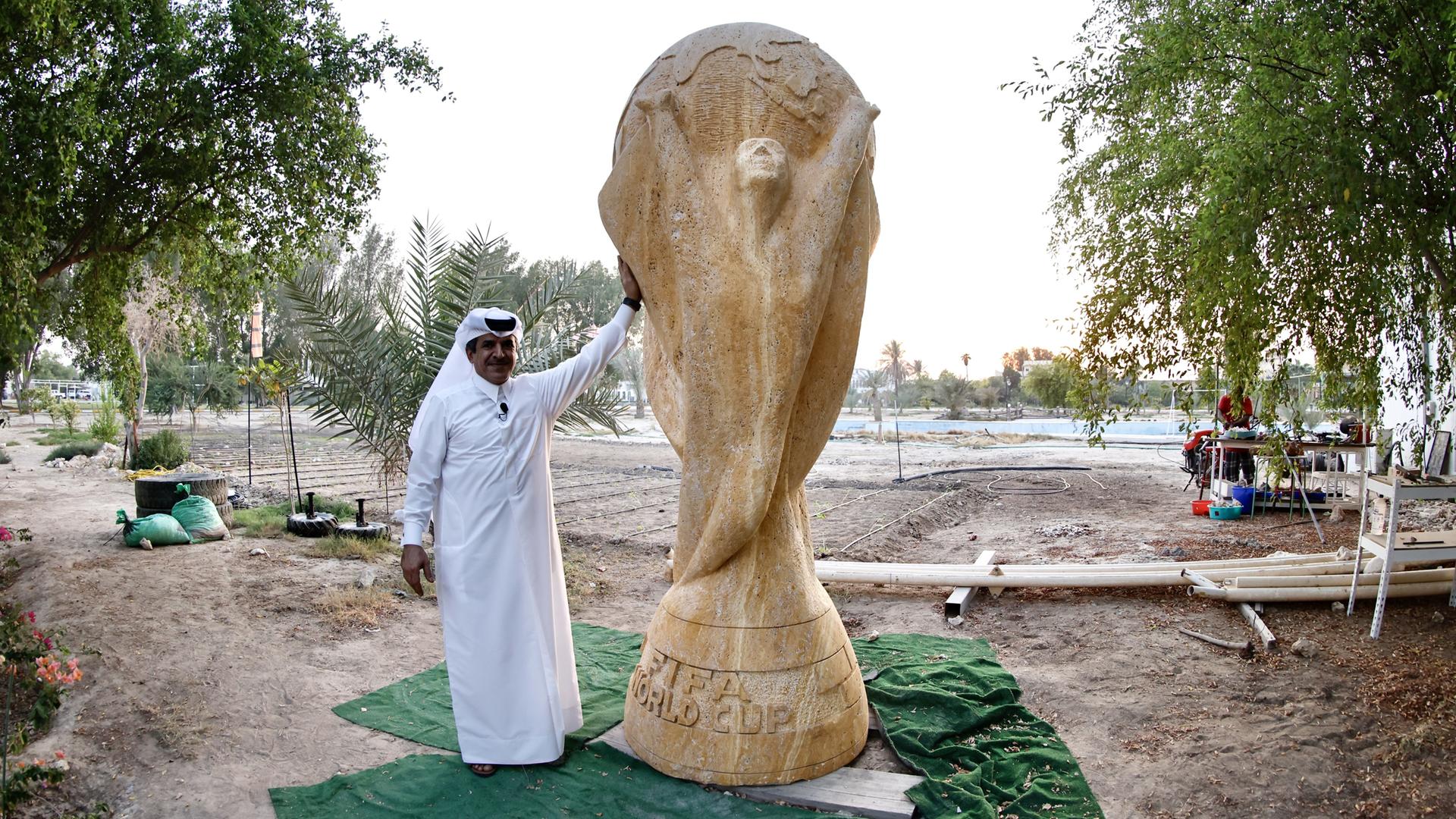 Ein Geschäftsmann in weißem Umhang posiert neben einer beinahe drei Meter hohen Skulptur des FIFA-Pokals aus sandfarbenem Travertin.