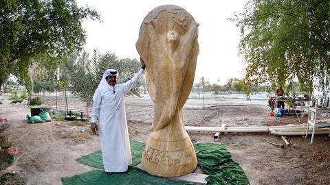 Ein Geschäftsmann in weißem Umhang posiert neben einer beinahe drei Meter hohen Skulptur des FIFA-Pokals aus sandfarbenem Travertin.