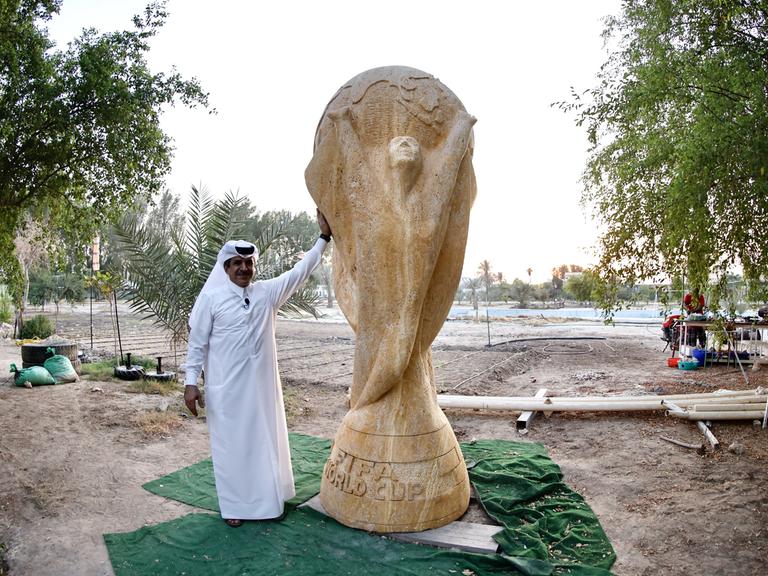 Ein Geschäftsmann in weißem Umhang posiert neben einer beinahe drei Meter hohen Skulptur des FIFA-Pokals aus sandfarbenem Travertin.