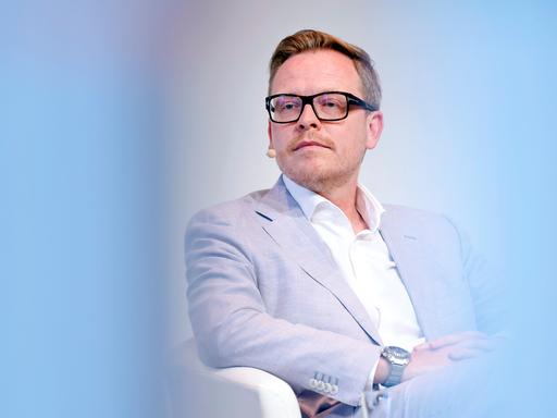 Porträt des Philosophen Markus Gabriel in einer Phil.Cologne Veranstaltung. Um ihn herum ist der Vordergrund hellblau verschwommen. Er trägt einen hellblauen Anzug und eine schwarze Brille.