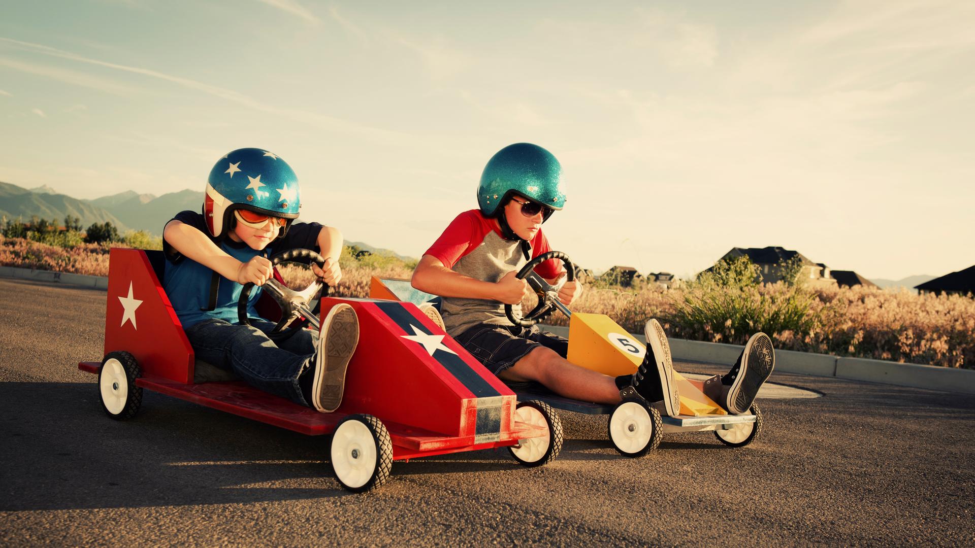 Zwei Kinder in Seifenkisten und coolen Helmen nebeneinander auf der Straße - das Rennen scheint gleich loszugehen