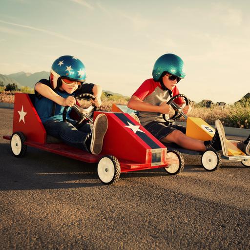 Zwei Kinder in Seifenkisten und coolen Helmen nebeneinander auf der Straße - das Rennen scheint gleich loszugehen