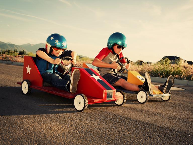 Zwei Kinder in Seifenkisten und coolen Helmen nebeneinander auf der Straße - das Rennen scheint gleich loszugehen